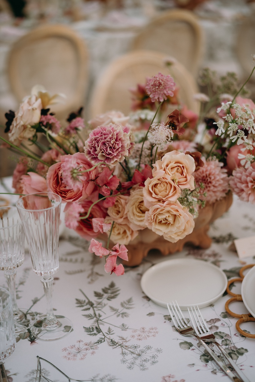 dettaglio centrotavola floreale dai colori desaturati con rose, gerbere, dalie e scabiosa per cena di matrimonio all'aperto su tovaglia con stampa botanica